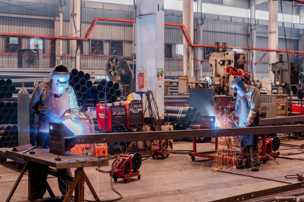 Two welders wearing protective gear working in a busy industrial factory workshop.